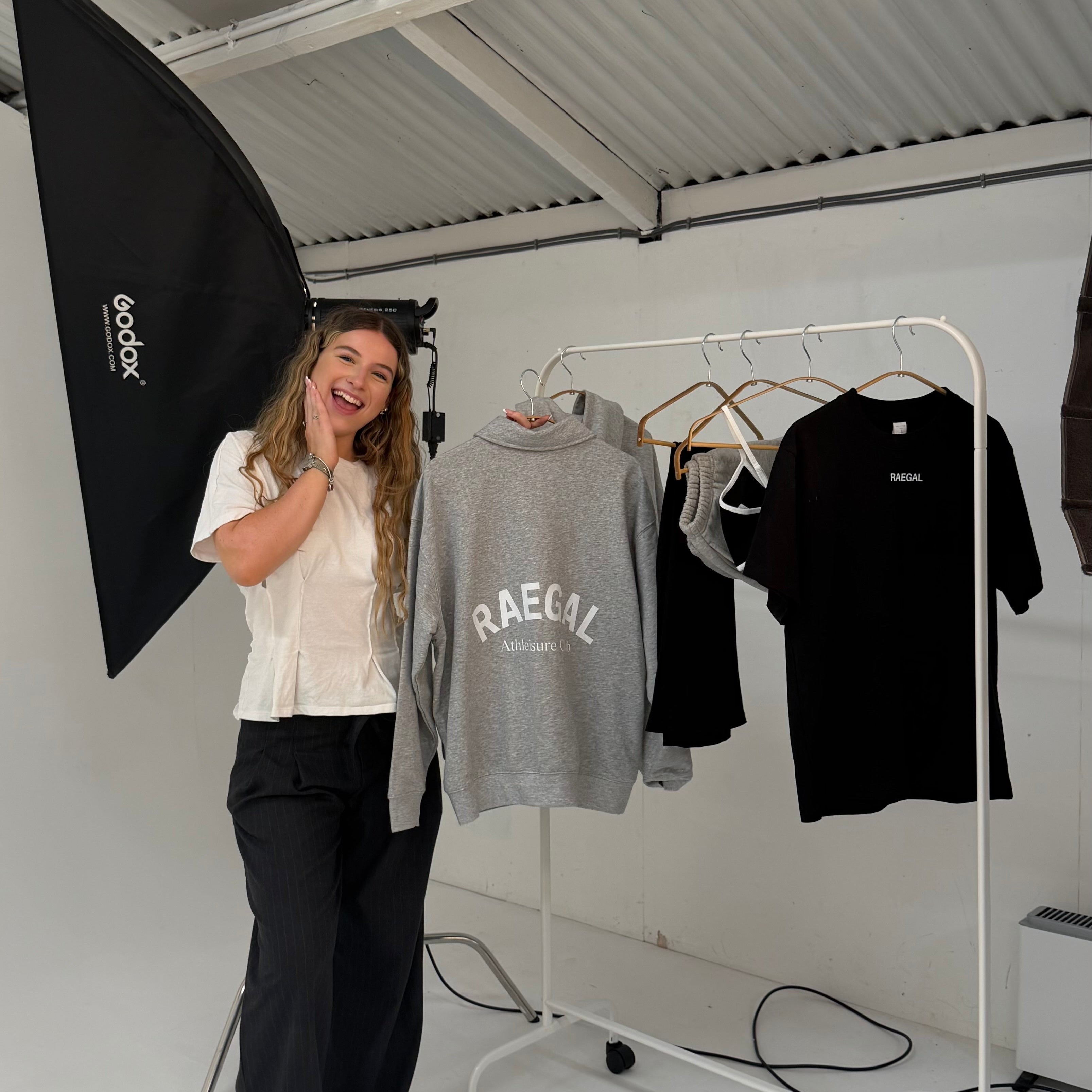 A woman stands in a studio next to a clothing rack holding a gray sweatshirt with 'RAEGAL' printed on the front, alongside black t-shirts. She is holding a black softbox light in a white studio space with a corrugated metal ceiling.